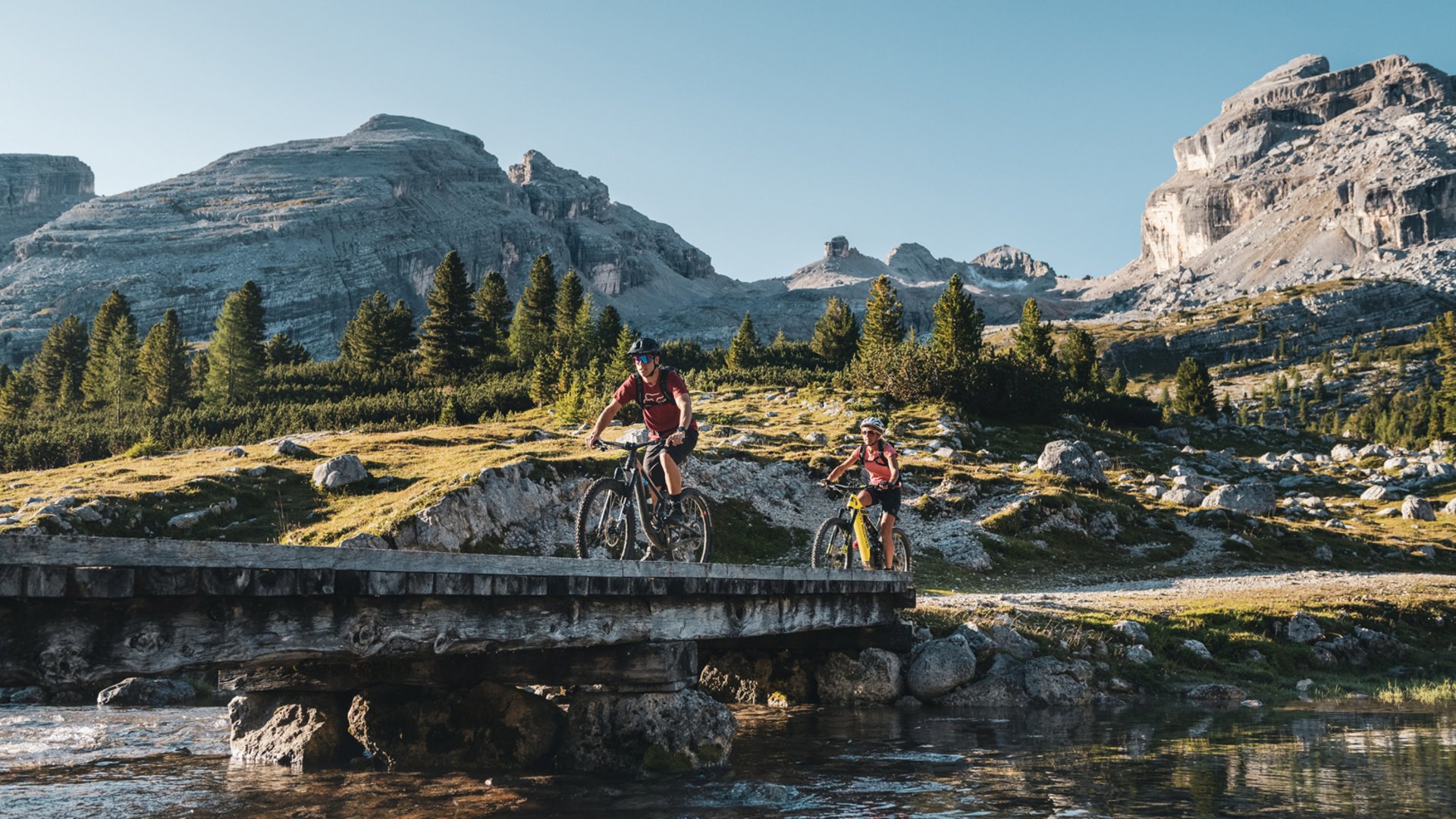 Excelsior: noleggio bici a San Vigilio di Marebbe Due ciclisti in mountain bike attraversano un ponte tra le montagne sotto un cielo sereno