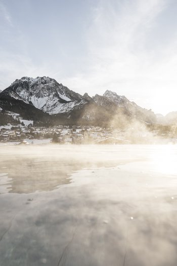 Hotel in den Dolomiten für adults only Dampf über See mit verschneiten Bergen und Dorf im Hintergrund bei Sonnenuntergang