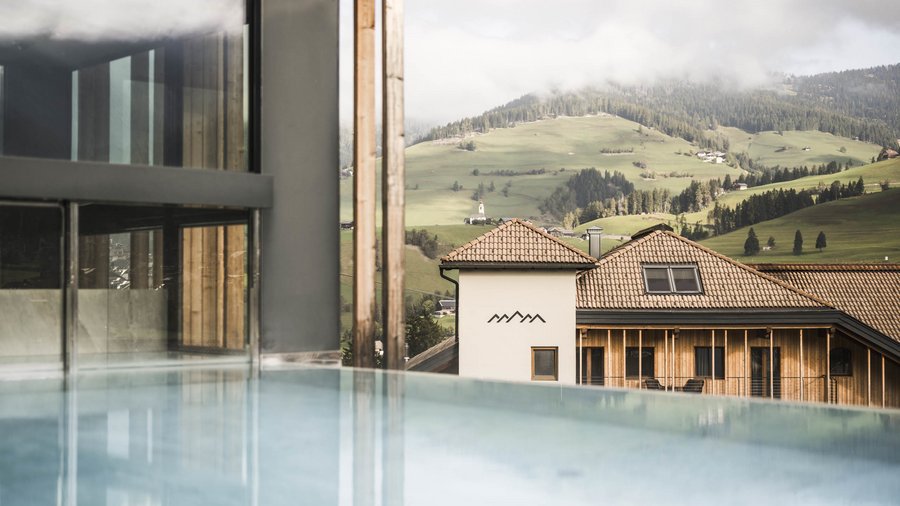 Excelsior: Wanderhotel in den Dolomiten Infinity-Pool mit Blick auf ein Haus und grüne Hügel in den Bergen