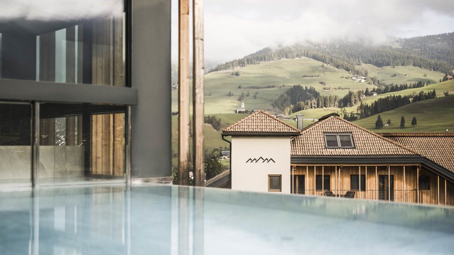 Excelsior: Wanderhotel in den Dolomiten Infinity-Pool mit Blick auf ein Haus und grüne Hügel in den Bergen