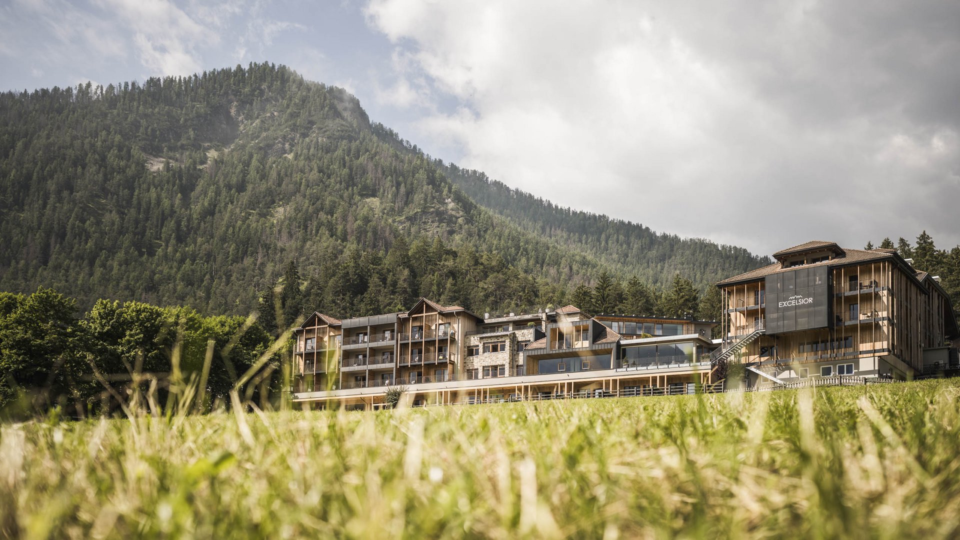 Luxushotel in den Dolomiten | Excelsior in St. Vigil Hotelgebäude vor bewaldetem Berg und bewölktem Himmel