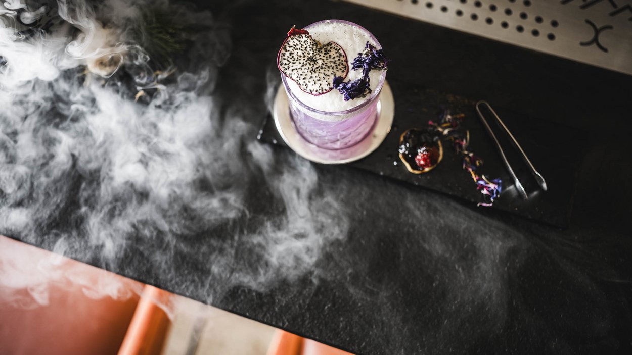 Sustainable hotels in South Tyrol? The Excelsior! Purple cocktail with foam and decorations on black table with smoke