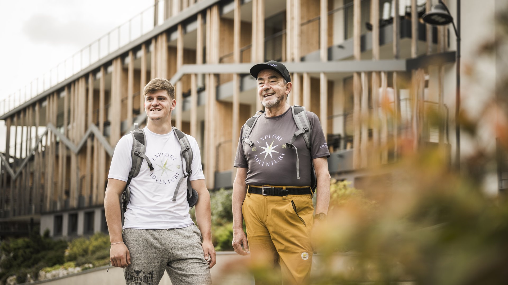 Excelsior: Wanderhotel in den Dolomiten Junger und älterer Mann mit Rucksäcken vor modernem Holzgebäude