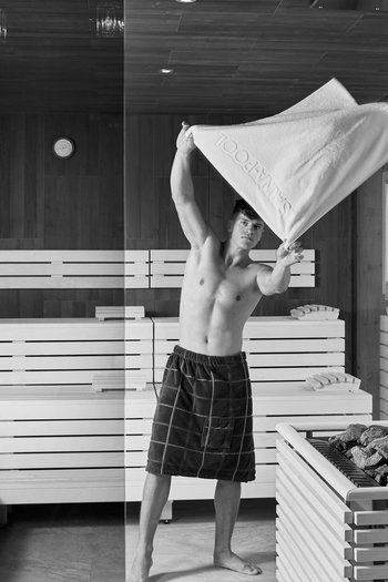 Benvenuti nel vostro hotel al Plan de Corones! Uomo che agita un asciugamano in una sauna moderna con pareti in legno e pietre.