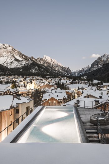 Hotel in den Dolomiten für adults only Schneebedeckte Bergstadt mit beleuchtetem Pool auf dem Dach bei Abenddämmerung