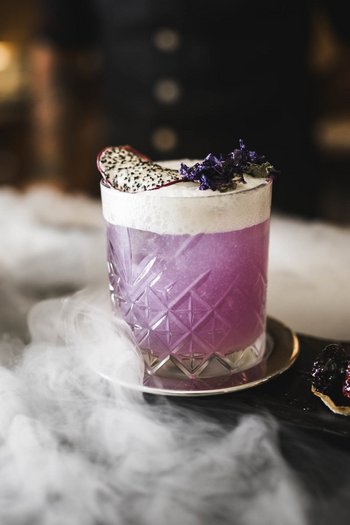 Das Gourmethotel in Südtirol Lila Cocktail mit Schaum und Früchten auf einem Tablett mit Nebel
