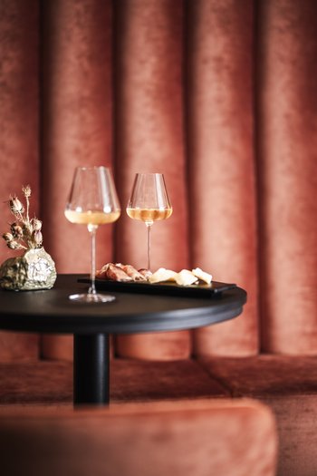 Il vostro hotel gourmet in Alto Adige Due calici di vino e piatto con affettati su tavolo nero davanti a divano rosso in velluto