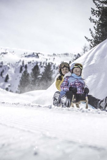 Hotel am Kronplatz direkt an der Piste Zwei Kinder rodeln zusammen auf einem Schneehügel mit verschneiten Bergen im Hintergrund
