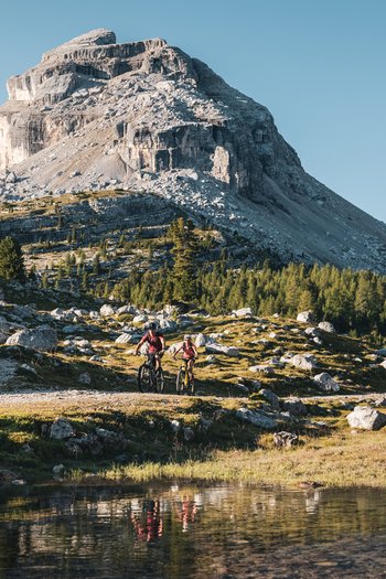 San Vigilio di Marebbe: escursioni per famiglie Due ciclisti su un sentiero di montagna con una grande roccia e un lago sullo sfondo