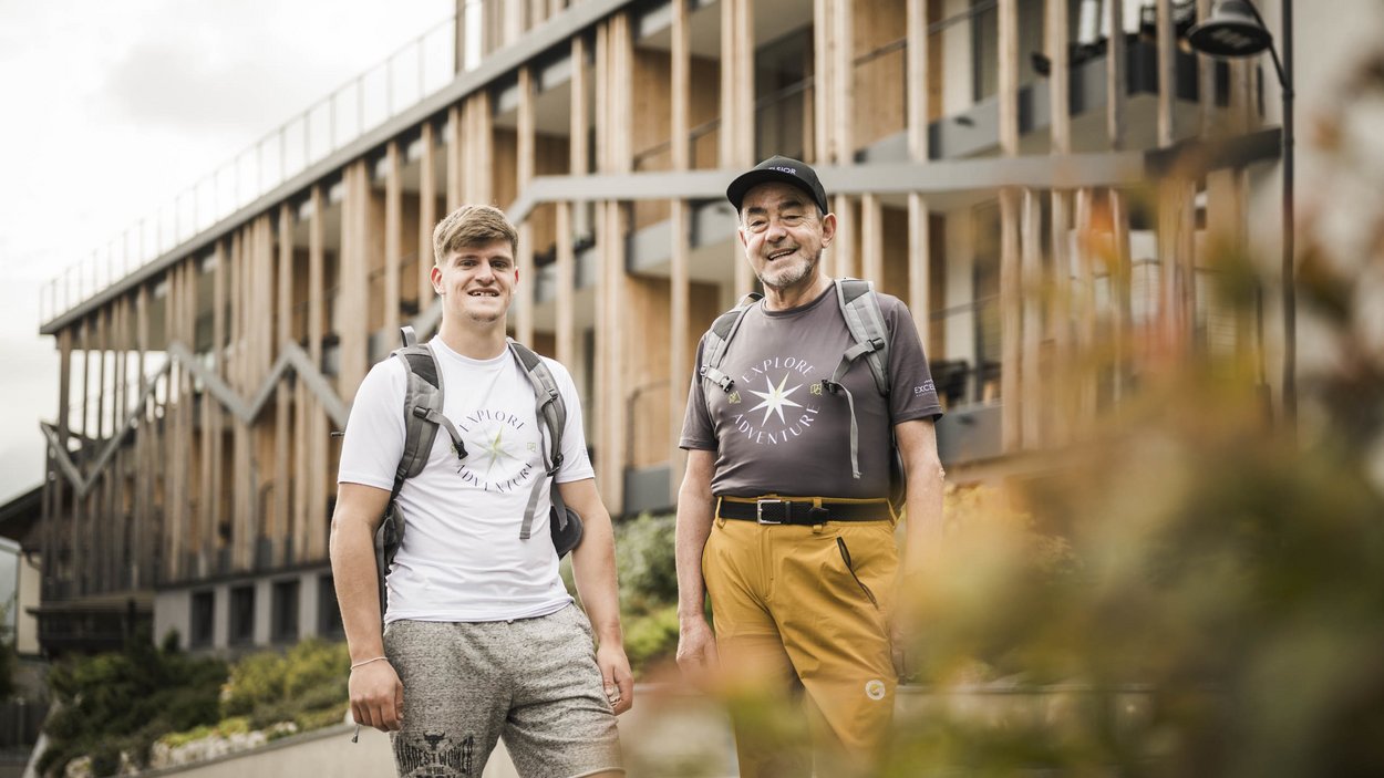 Excelsior: Wanderhotel in den Dolomiten Zwei Männer mit Rucksäcken stehen vor modernem Gebäude im Freien