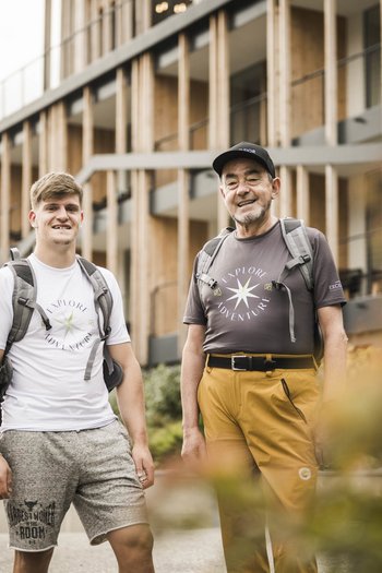 Excelsior: Wanderhotel in den Dolomiten Zwei Männer mit Rucksäcken stehen vor modernem Gebäude im Freien