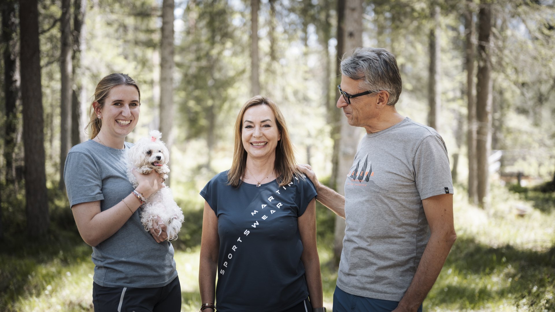 Drei lächelnde Personen im Wald, eine hält einen kleinen weißen Hund