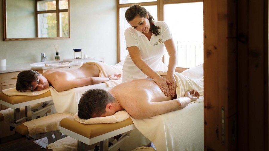Hotel adults only in Alto Adige: l’Excelsior! Donna massaggia schiena di uomo e donna sdraiati su lettini spa in stanza luminosa.
