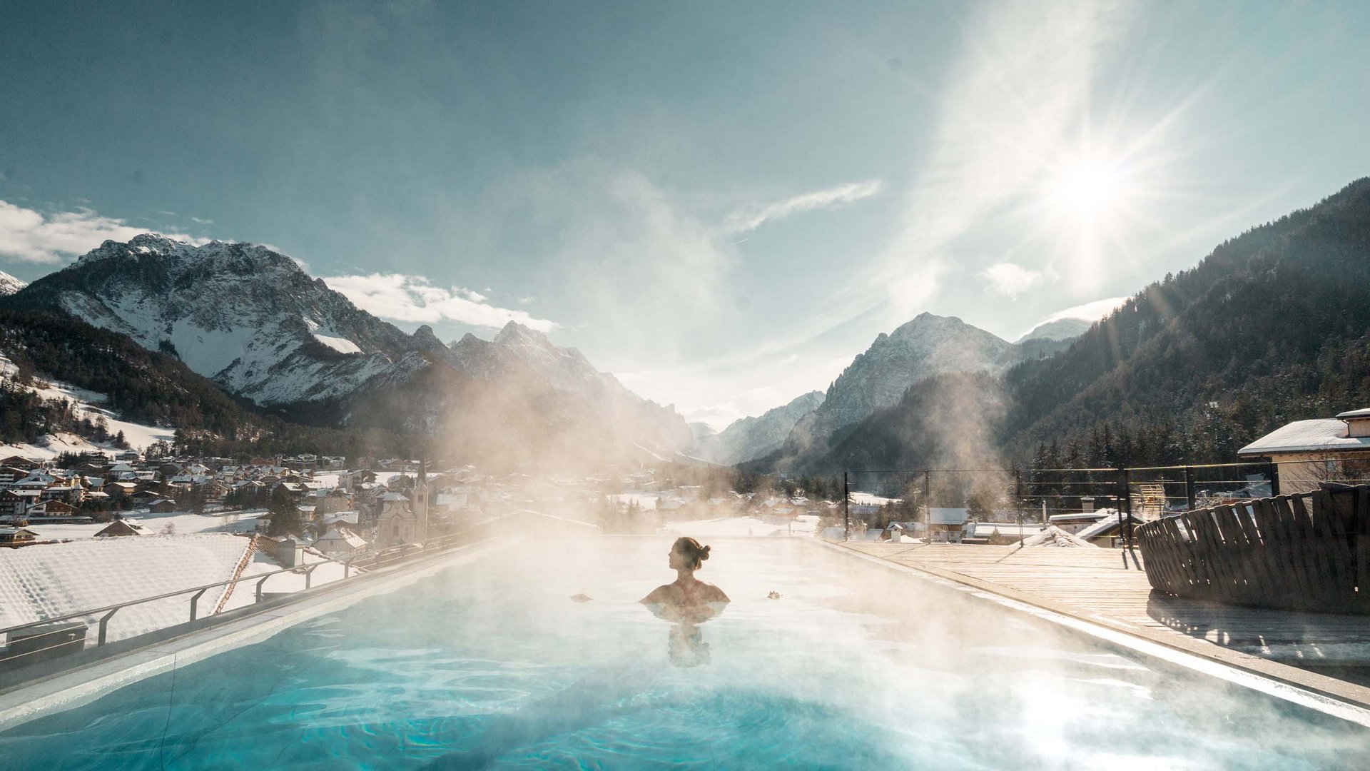 Events: Excelsior Dolomites Life Resort Person in heated outdoor pool with view of snowy mountains and sunny sky
