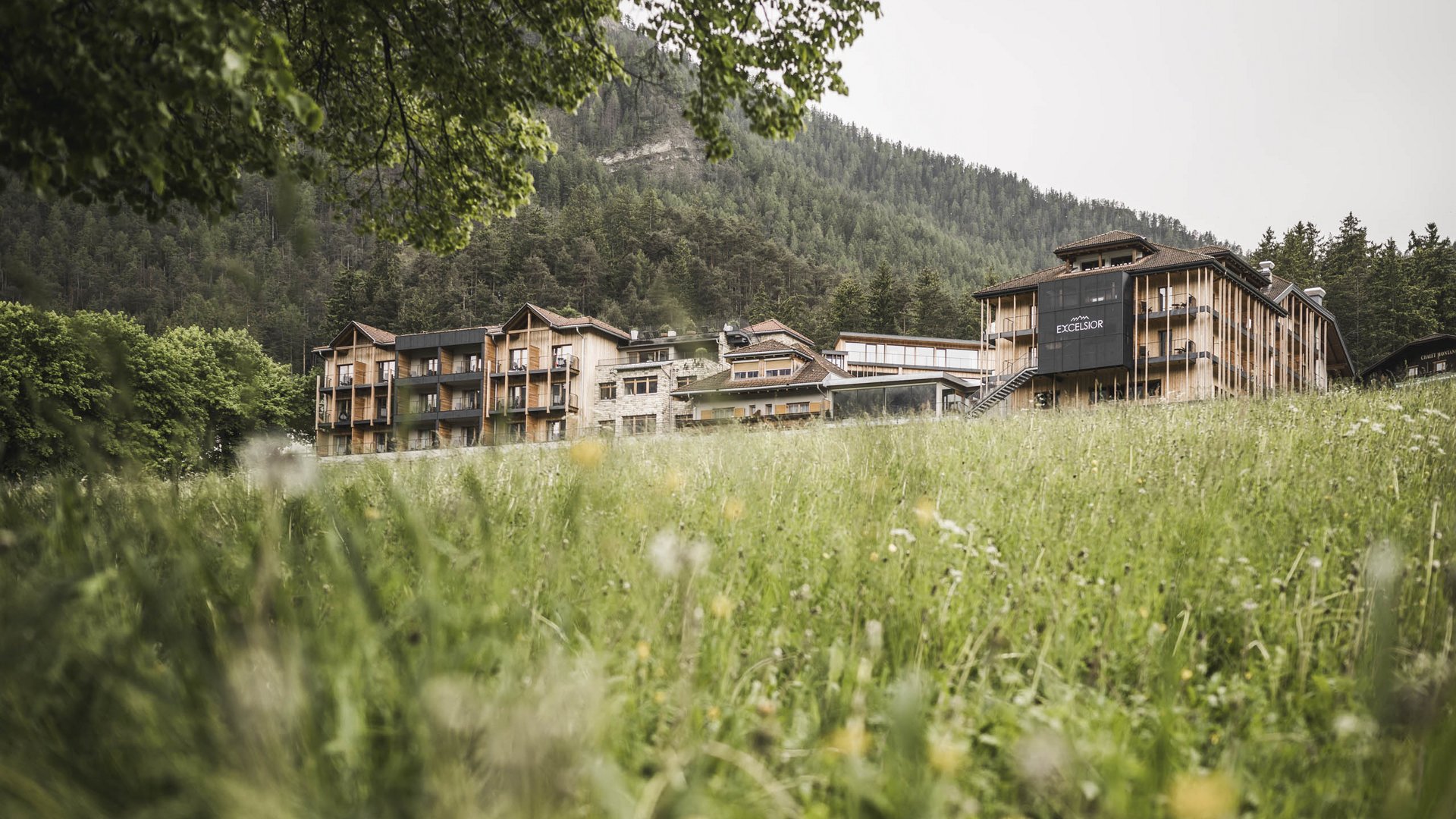 Wellnesshotel in den Dolomiten Hotelgebäude in grüner Wiesenlandschaft vor bewaldetem Berg