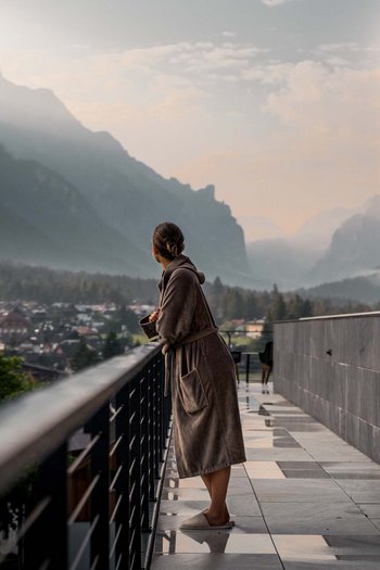 Hotel adults only in Alto Adige: l’Excelsior! Donna in accappatoio su balcone con vista su paesaggio montano e villaggio