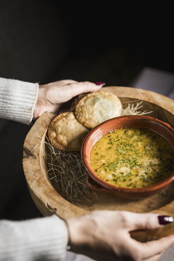 Wellnesshotel in Südtirol: Auf in den Traumurlaub! Hände halten ein Holztablett mit einer Suppe und zwei goldbraunen Teigtaschen