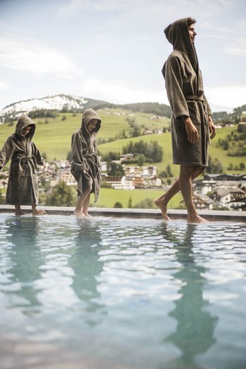 Excelsior: hotel wellness in Alto Adige per famiglie Uomo e due bambini in accappatoi vicino alla piscina con vista sulle montagne