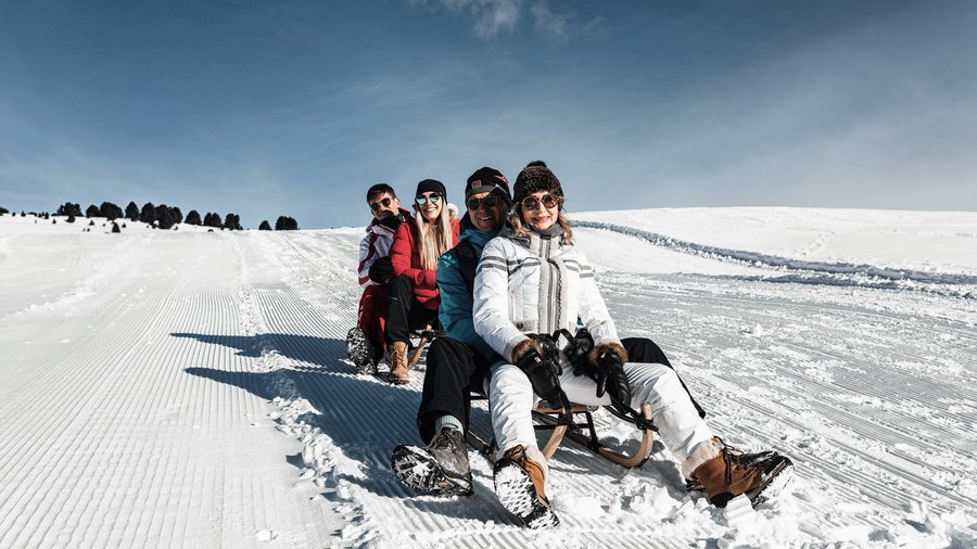 Mountain hotel in the Dolomites/South Tyrol Four people sledding together down a snowy hill under clear sky