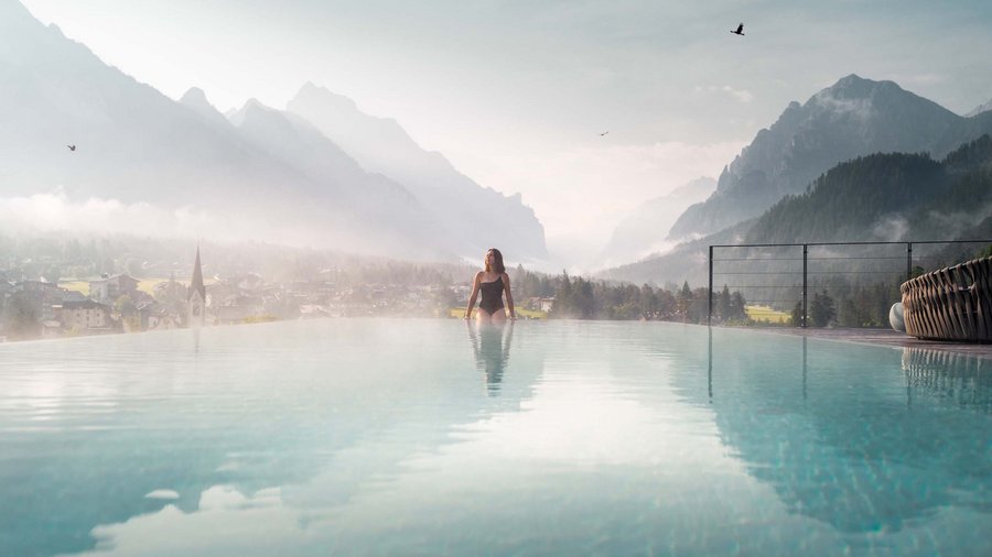 Luxushotel in den Dolomiten | Excelsior in St. Vigil Frau im Infinity-Pool mit Bergblick an nebligem Morgen