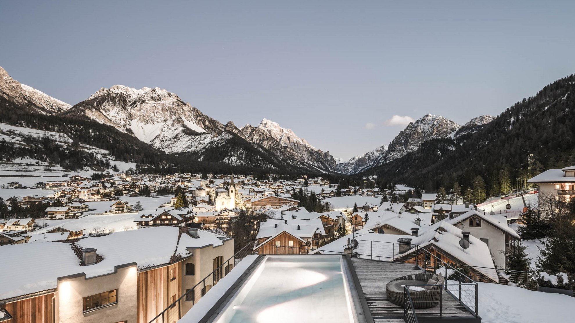 Events: Excelsior Dolomites Life Resort Snow-covered village in the mountains with lit-up village center at dusk