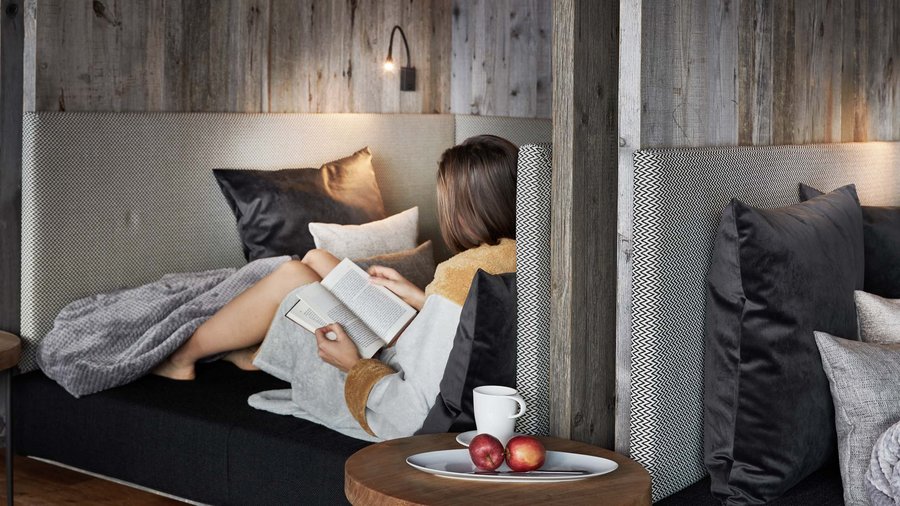 Wellnesshotel in den Dolomiten Frau entspannt auf Couch mit Buch und Obst auf Beistelltisch