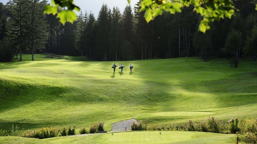 Hotel in South Tyrol with guided hikes Three golfers carrying bags on green golf course with forest in background