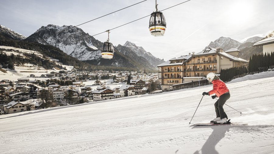 Hotel am Kronplatz direkt an der Piste Skifahrer fährt den Hang hinunter mit Seilbahnen und Dorf im Hintergrund