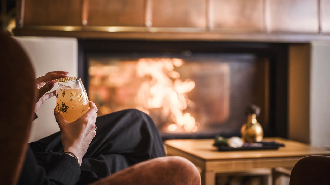 Il vostro hotel gourmet in Alto Adige Persona che si rilassa con una bevanda davanti a un camino acceso
