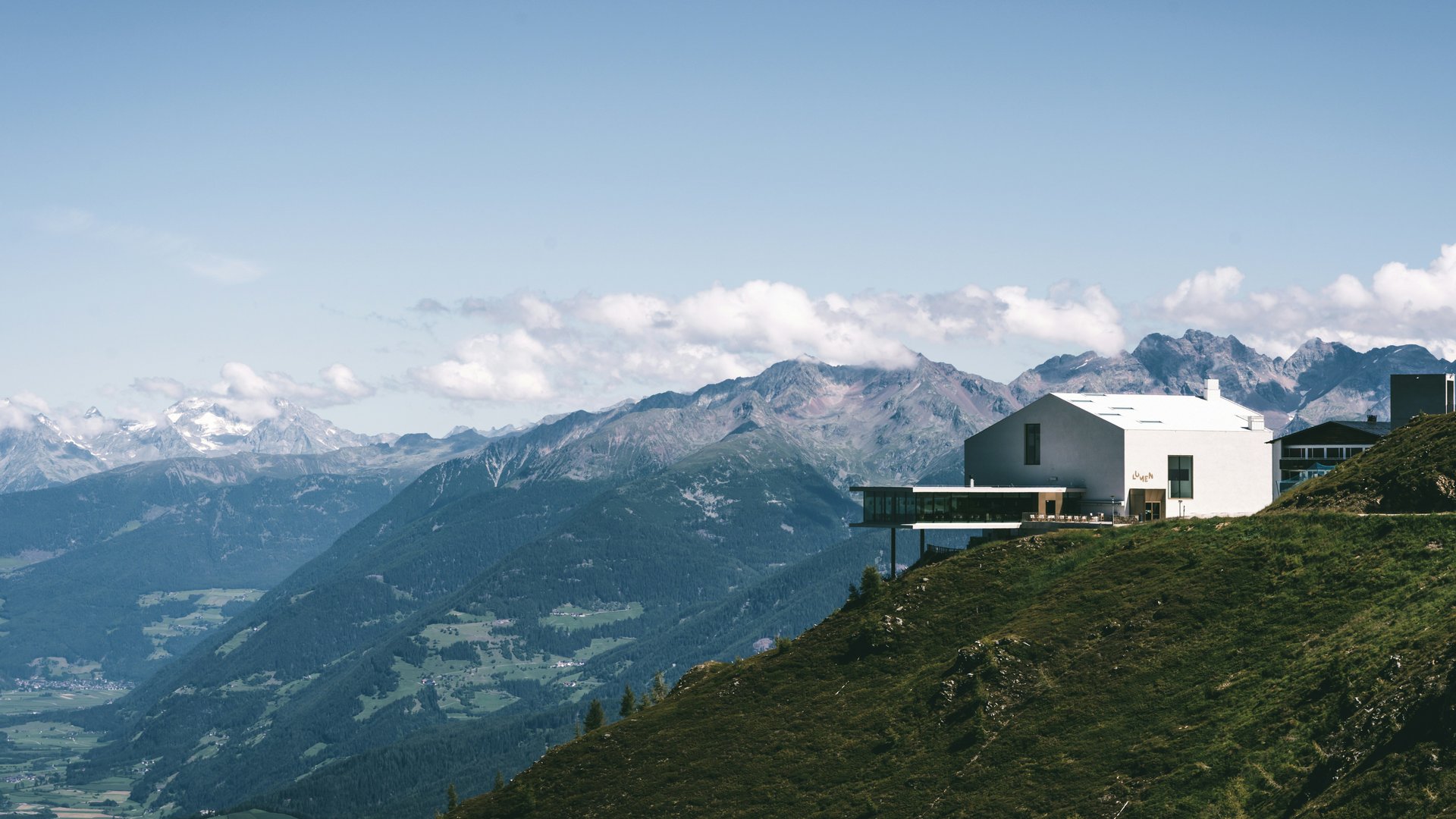 Haus auf grünem Hang mit Bergpanorama und blauem Himmel