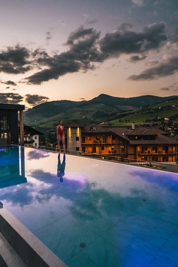 Hotel con infinity pool in Alto Adige: l’Excelsior Donna in piedi al bordo di una piscina a sfioro illuminata con vista sulle montagne al tramonto