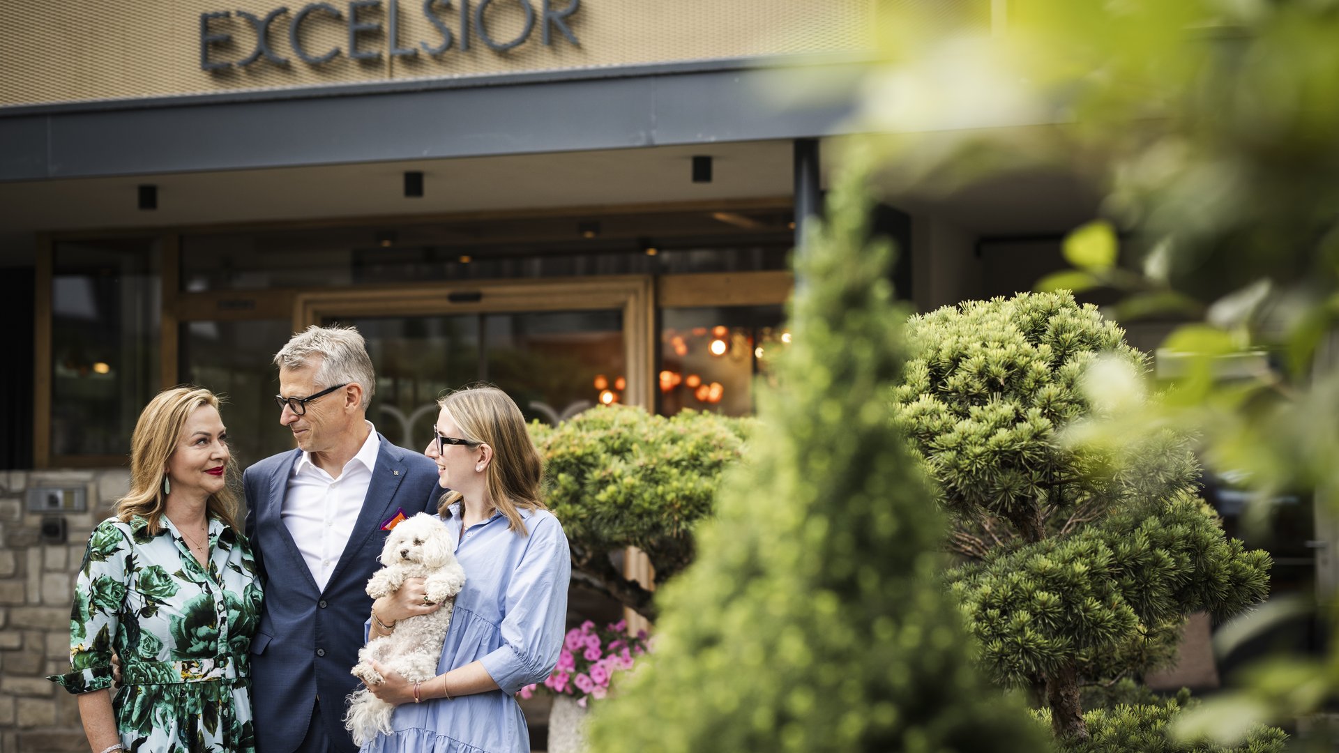 Dolomiten-Hotel für Bergmenschen Drei Personen mit Hund vor dem Excelsior Hotel mit grüner Pflanzenkulisse