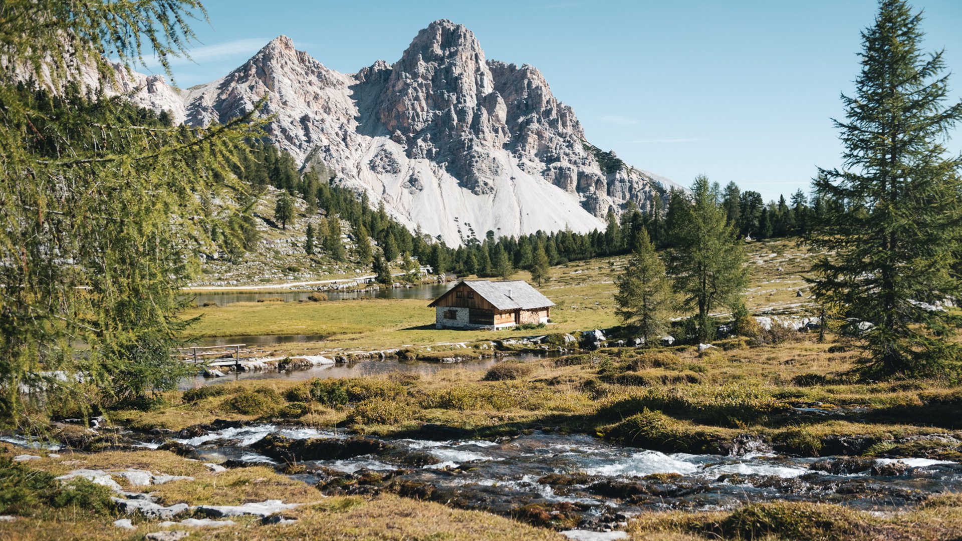 Nei dintorni di San Vigilio: escursioni indimenticabili Baita alpina davanti a montagna e pini con ruscello in primo piano