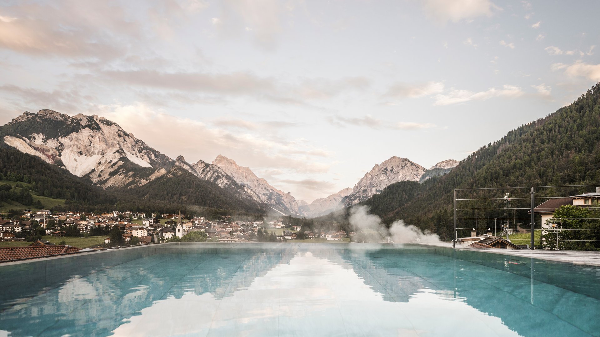 Hotel con infinity pool in Alto Adige: l’Excelsior Piscina a sfioro con vista sulle Alpi e villaggio al crepuscolo