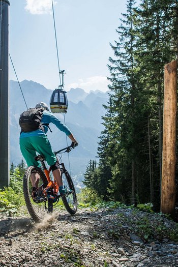 Eines der Bike-Hotels in Südtirol Mountainbiker startet auf Trail Piz de Plaies im Wald mit Bergseilbahn im Hintergrund