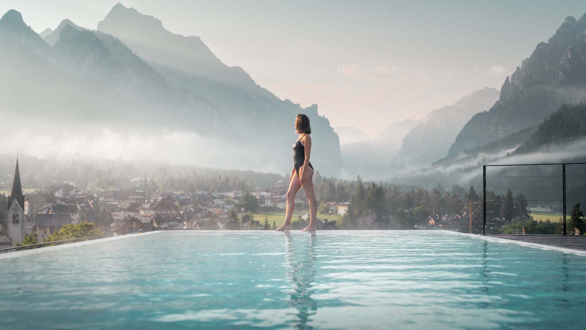 Hotel in the Dolomites for adults only Woman in swimsuit standing by pool with mountain and village view