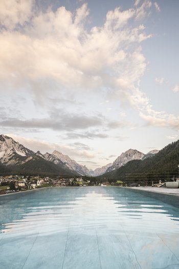 Wellnesshotel in den Dolomiten Infinity-Pool mit Blick auf Berge und Dorf unter bewölktem Himmel