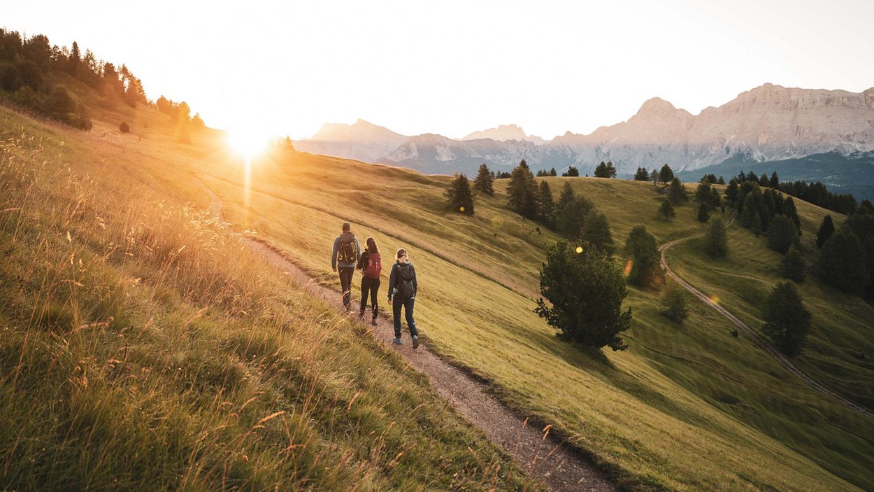 Excelsior: Wanderhotel in den Dolomiten Drei Wanderer auf Bergpfad bei Sonnenuntergang mit Alpen im Hintergrund