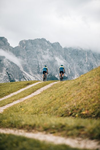 Excelsior: Wanderhotel in den Dolomiten Zwei Radfahrer auf einem Bergweg mit nebligen Gipfeln im Hintergrund