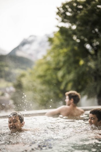 Familienurlaub in den Dolomiten – Natur, Erholung & Abenteuer Zwei Kinder spielen und plantschen im Pool, im Hintergrund sitzt ein Erwachsener im Wasser