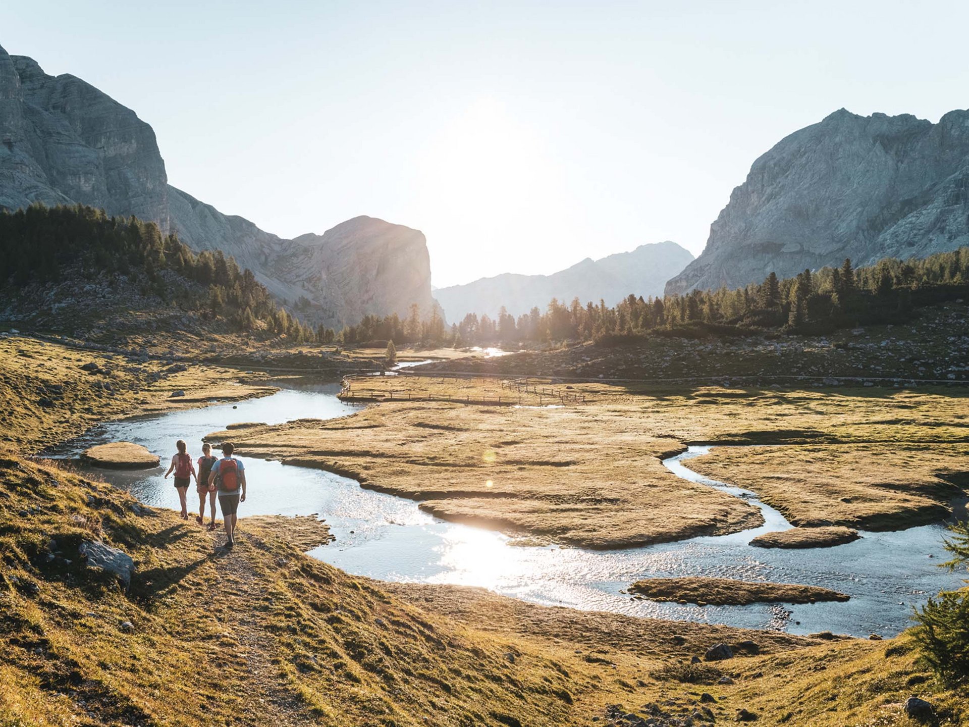 Kurzurlaub in Südtirol? Im Excelsior Dolomites Life Resort Drei Wanderer gehen an einem Fluss in einer sonnigen Berglandschaft
