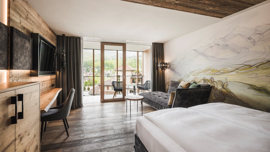 Wohnen am Kronplatz: Hotel an der Piste Modernes Hotelzimmer mit Holzboden, Sofa und Balkon mit Bergblick