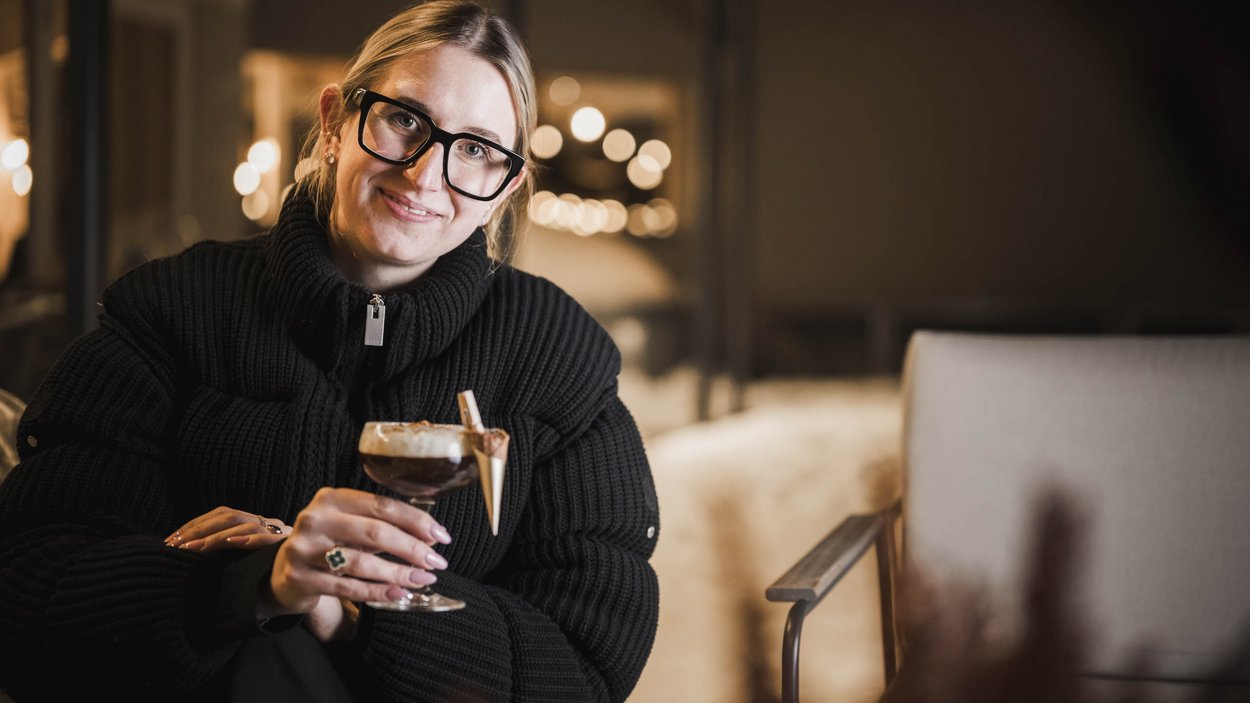 #myexcelsior Sunday News 09.02.2025 Donna con occhiali grandi e maglione nero tiene un bicchiere da dessert in ambiente accogliente