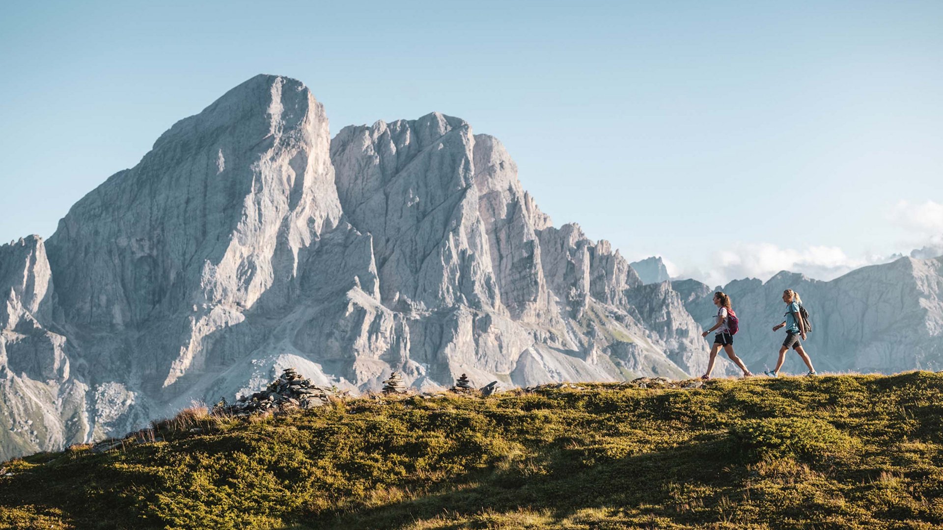 Pietre Preziose Due escursionisti camminano su una collina erbosa con montagne sullo sfondo