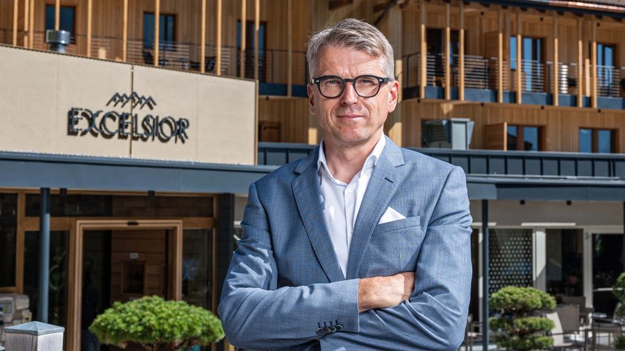 Dolomites hotel for mountain people Man in suit standing in front of Excelsior hotel with wooden facade and mountains behind