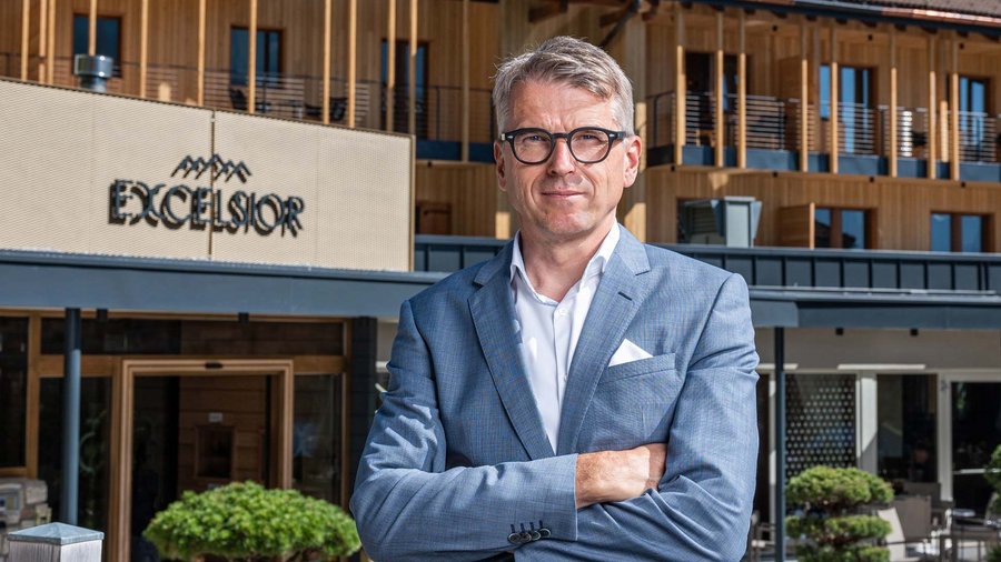 Dolomiten-Hotel für Bergmenschen Mann in Anzug vor Hotel Excelsior mit Holzfassade und Bergen im Hintergrund