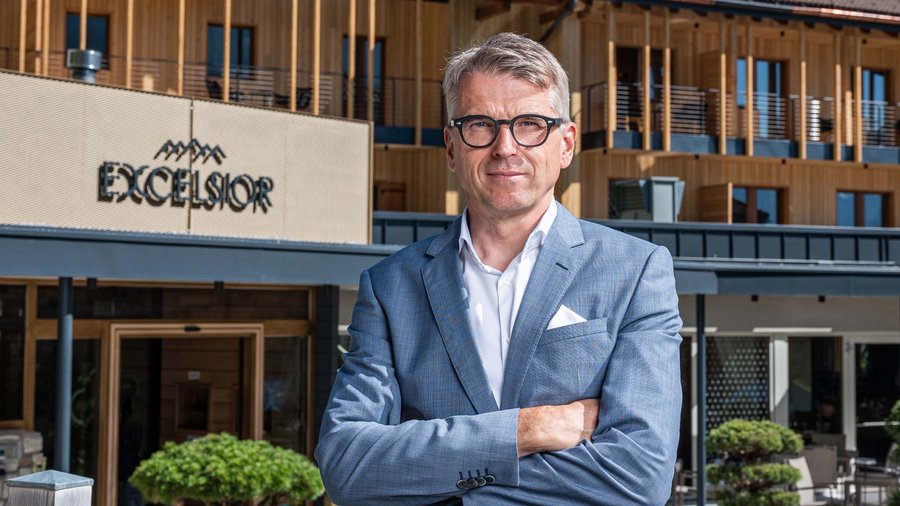 Dolomiten-Hotel für Bergmenschen Mann in Anzug vor Hotel Excelsior mit Holzfassade und Bergen im Hintergrund