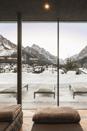 Hotel in den Dolomiten für adults only Blick durch große Fenster auf schneebedeckte Berge und Liegestühle im Innen- und Außenbereich