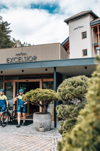 Excelsior: Wanderhotel in den Dolomiten Zwei Radfahrer vor dem Eingang des Hotels Excelsior mit Bäumen und Bergen
