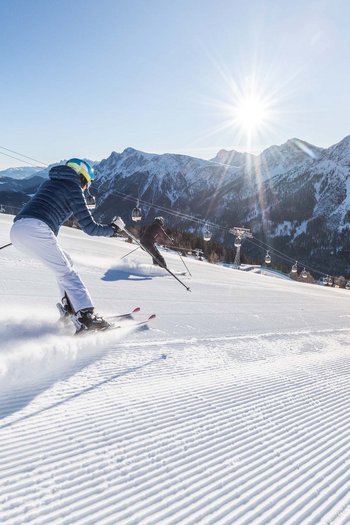 Around the hotel on the slopes in South Tyrol Two skiers on freshly groomed slope with mountains and sun in background